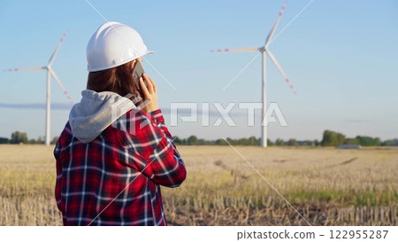 Woman engineer is talking on a smartphone in a field with wind turbines as the sun sets. Concept of clean energy and engineering audit Woman engineer is talking on a smartphone in a field with wind turbines as the sun sets. Concept of clean energy and engineering audit 122955287