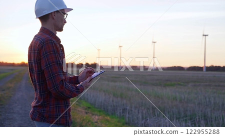 Man engineer wearing a white protective helmet is taking notes with a clipboard in a field with wind turbines, as the sun sets. Clean energy and engineering concept 122955288