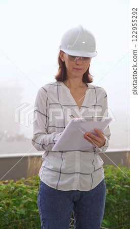 Woman a construction engineer wearing white checked blouse and hard hat is making notes on a clipboard while inspecting a building site in foggy weather, front vertical view. Architecture concept 122955292