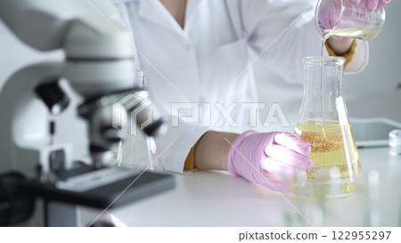 A scientist, wearing a lab coat and pink protective gloves, is pouring a yellow oily liquid from one beaker to another near microscope in laboratory, close up. Medicine and science 122955297