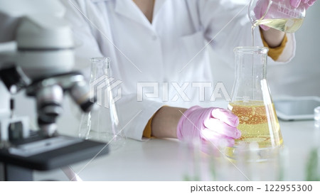A scientist, wearing a lab coat and pink protective gloves, is pouring a yellow oily liquid from one beaker to another near microscope in laboratory, close up. Medicine and science 122955300