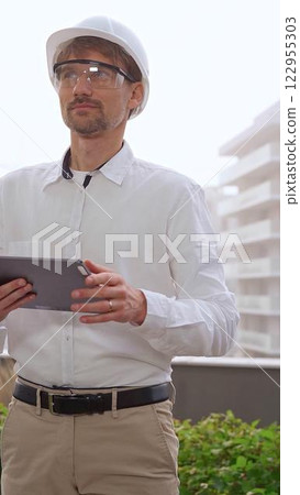 Man a construction engineer or architect, wearing a white shirt and helmet, is using a digital tablet while inspecting a building site in foggy weather, front view. Architecture and engineering 122955303