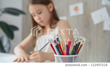 A close-up of colored pencils on the desk, with a creative young girl drawing in the background A close-up of colored pencils on the desk, with a creative young girl drawing in the background 122955328