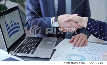 Two business people professionals shaking hands in office setting, sealing a deal with laptop and financial graphs visible. Business handshake over laptop and reports Two business people professionals shaking hands in office setting, sealing a deal with laptop and financial graphs visible. Business handshake over laptop and reports 122955388
