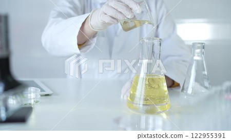 A scientist, wearing a lab coat and white protective gloves, is pouring a yellow oily liquid from one beaker to another in laboratory, close up. Medicine and science concept A scientist, wearing a lab coat and white protective gloves, is pouring a yellow oily liquid from one beaker to another in laboratory, close up. Medicine and science concept 122955391