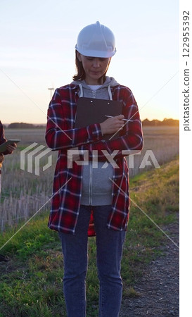 Woman engineer is standing straight and taking notes with a clipboard in a field with wind turbines, as the sun sets. Clean energy and engineering concept Woman engineer is standing straight and taking notes with a clipboard in a field with wind turbines, as the sun sets. Clean energy and engineering concept 122955392