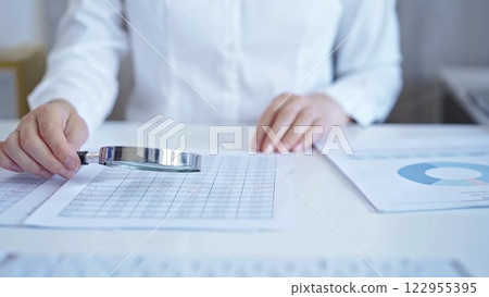 Businesswoman with white blues is analyzing financial data on a spreadsheet using a magnifying glass. Audit and taxes 122955395