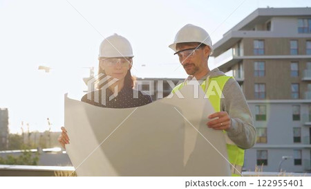 Two architects wearing safety hard hats are holding blueprint and discussing something on construction site at sunrise. Architect and teamwork concept Two architects wearing safety hard hats are holding blueprint and discussing something on construction site at sunrise. Architect and teamwork concept 122955401