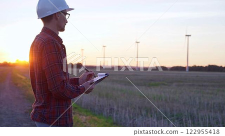Man engineer wearing a white protective helmet is taking notes with a clipboard in a field with wind turbines, as the sun sets. Clean energy and engineering concept Man engineer wearing a white protective helmet is taking notes with a clipboard in a field with wind turbines, as the sun sets. Clean energy and engineering concept 122955418