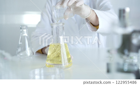 A scientist, wearing a lab coat and white protective gloves, is pouring a yellow oily liquid from one beaker to another in laboratory, close up. Medicine and science concept A scientist, wearing a lab coat and white protective gloves, is pouring a yellow oily liquid from one beaker to another in laboratory, close up. Medicine and science concept 122955486