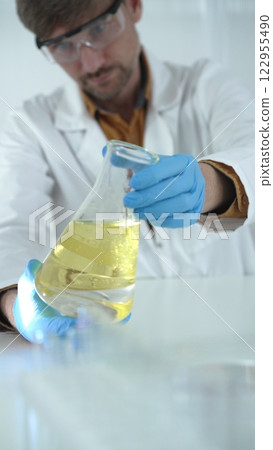 Man scientist, wearing blue gloves and protective glasses, is holding a yellow chemical solution inside Erlenmeyer flask in a laboratory, vertical portrait view. Science and medicine 122955490