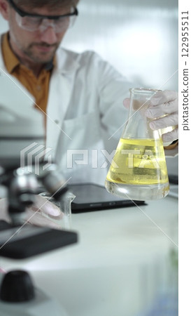 Man scientist wearing white protective gloves, and glasses is holding a yellow chemical solution inside an Erlenmeyer flask in a laboratory, vertical portrait. Science and medicine 122955511