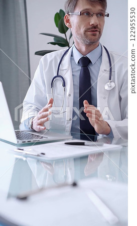 Doctor consulting patient. Male physician is gesturing above a clipboard while talking with a woman in a clinic, close up Doctor consulting patient. Male physician is gesturing above a clipboard while talking with a woman in a clinic, close up 122955530