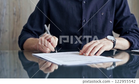 Businessman in dark blue t-shirt is signing contract at desk. Close-up of a male executive signing a legal document on a glass desk. Business people concept Businessman in dark blue t-shirt is signing contract at desk. Close-up of a male executive signing a legal document on a glass desk. Business people concept 122955542
