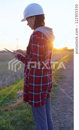 Woman engineer wearing a white protective helmet is taking notes with a clipboard in a field with wind turbines, as the sun sets. Clean energy and engineering concept 122955550