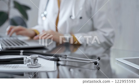 Stethoscope and clipboard with medication history records are on a glass desk behind a physician using a laptop computer. Medicine concept 122955593