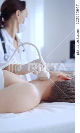 Doctor woman with white medical gloves performing neck ultrasound exam on male patient. Thyroid gland examination. Medicine concept 122955647