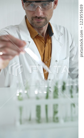 A male science researcher, wearing a lab coat and protective glasses, drips liquid from a pipette into a test tube containing a green plant. His face is focused, captured in a vertical portrait 122955689