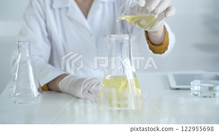 A scientist, wearing a lab coat and white protective gloves, is pouring a yellow oily liquid from one beaker to another in laboratory, close up. Medicine and science concept A scientist, wearing a lab coat and white protective gloves, is pouring a yellow oily liquid from one beaker to another in laboratory, close up. Medicine and science concept 122955698