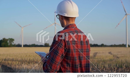 Man engineer at work is wearing a white protective helmet and taking notes with a tablet computer in a field with wind turbines, as the sun sets. Clean energy concept 122955720