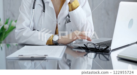 Thinking doctor woman is reviewing medical records on laptop. Close-up of a healthcare unknown professional analyzing patient information on a computer. Medicine 122955724
