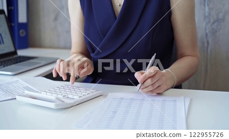 Businesswoman with dark blue dress is using white calculator and taking notes in her office. Taxes and audit in business 122955726