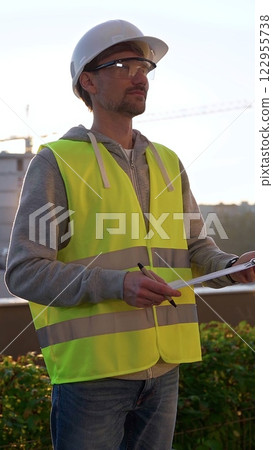 Man engineer with white helmet and safety vest is taking notes on a clipboard while inspecting a construction site at sunrise in early morning, vertical front view. Architecture and engineering 122955738