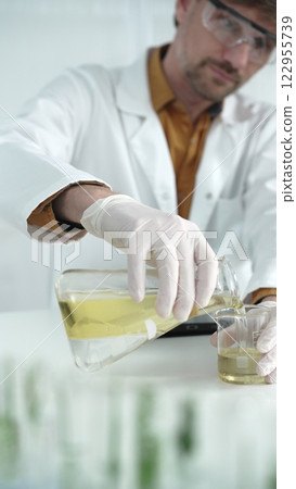 Man scientist with lab coat, white gloves, and protective glasses, is pouring a yellow oily liquid from one beaker to another near microscope in a laboratory, vertical close-up view Man scientist with lab coat, white gloves, and protective glasses, is pouring a yellow oily liquid from one beaker to another near microscope in a laboratory, vertical close-up view 122955739