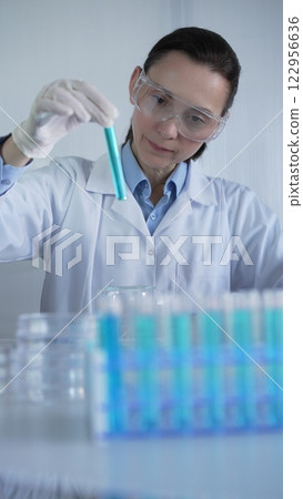 Female lab worker or scientist, wearing coat, blue medical gloves, and protective glasses is examining test tube with blue liquid in modern laboratory. Low key lighting photo. Science and Medicine Female lab worker or scientist, wearing coat, blue medical gloves, and protective glasses is examining test tube with blue liquid in modern laboratory. Low key lighting photo. Science and Medicine 122956636