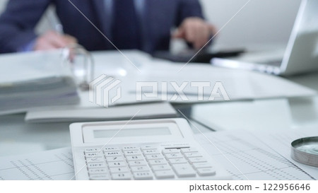 Magnifying glass, calculator, pen, laptop and documents are lying on accountant's desk. Businessman wearing blue suit is working with documents on the background. Business people and audit concept Magnifying glass, calculator, pen, laptop and documents are lying on accountant's desk. Businessman wearing blue suit is working with documents on the background. Business people and audit concept 122956646