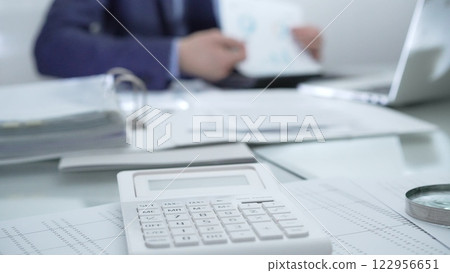 Calculator, pen, laptop and documents are lying on accountant's desk. Businessman wearing blue suit is working with documents on the background. Business people, audit concept Calculator, pen, laptop and documents are lying on accountant's desk. Businessman wearing blue suit is working with documents on the background. Business people, audit concept 122956651