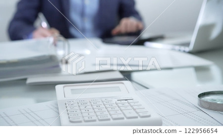 Magnifying glass, calculator, pen, laptop and documents are lying on accountant's desk. Businessman wearing blue suit is working with documents on the background. Business people and audit concept 122956654
