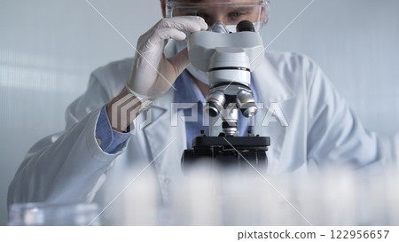 Man scientist researcher wearing white gloves, protective glasses and medical mask is using microscope in modern laboratory, portrait view. Low key lighting photo. Science and Medicine Man scientist researcher wearing white gloves, protective glasses and medical mask is using microscope in modern laboratory, portrait view. Low key lighting photo. Science and Medicine 122956657