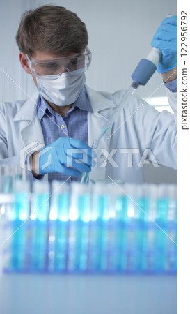 Male scientist researcher, wearing lab coat, blue medical gloves, mask and protective glasses is using micropipette filling test tubes in modern laboratory. Low key lighting. Science and Medicine 122956792