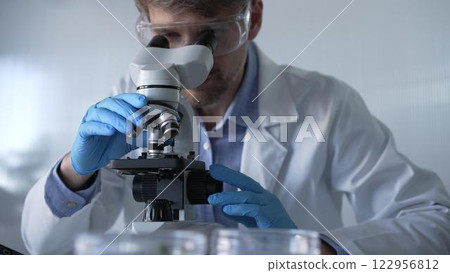 Male scientist researcher wearing blue gloves, protective glasses is using microscope in modern laboratory, portrait view. Low key lighting photo. Science and Medicine concepts 122956812