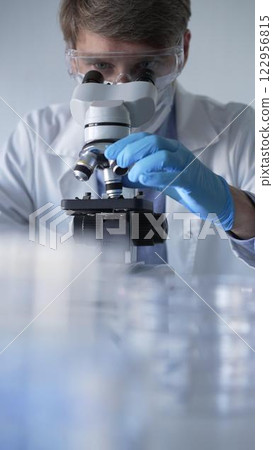 Man scientist researcher wearing blue gloves, protective glasses and medical mask is using microscope in modern laboratory, portrait view. Low key lighting photo. Science and Medicine concepts 122956815
