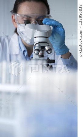 Woman scientist wearing blue gloves, protective glasses and medical mask is using microscope in modern laboratory setup. Low key lighting photo. Science and Medicine Woman scientist wearing blue gloves, protective glasses and medical mask is using microscope in modern laboratory setup. Low key lighting photo. Science and Medicine 122956838