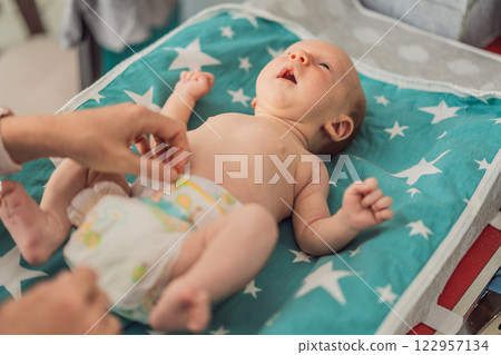 Young mother changing her baby diaper at home. Caring mom practicing hygiene routine with newborn child. Parenthood, baby care, and early motherhood concept 122957134