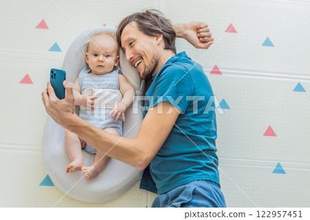 A 3-month-old baby lies safely in a cozy infant cocoon, secured with a special safety belt, while their father lies beside them during a video call. Modern parenting, bonding, and technology concept A 3-month-old baby lies safely in a cozy infant cocoon, secured with a special safety belt, while their father lies beside them during a video call. Modern parenting, bonding, and technology concept 122957451