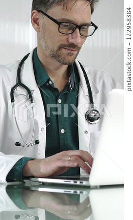 A male doctor in a green shirt under a medical coat is using technology in healthcare. Man is typing on a laptop on a glass desk in the clinic, vertical portrait view. Medicine A male doctor in a green shirt under a medical coat is using technology in healthcare. Man is typing on a laptop on a glass desk in the clinic, vertical portrait view. Medicine 122958384