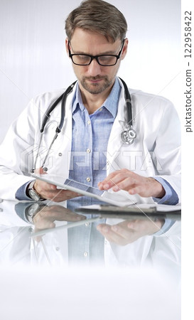 Doctor man in blue shirt under a white coat, glasses and stethoscope is using digital tablet in modern healthcare setting, interacting with patient data, vertical portrait view. Medicine and science Doctor man in blue shirt under a white coat, glasses and stethoscope is using digital tablet in modern healthcare setting, interacting with patient data, vertical portrait view. Medicine and science 122958422