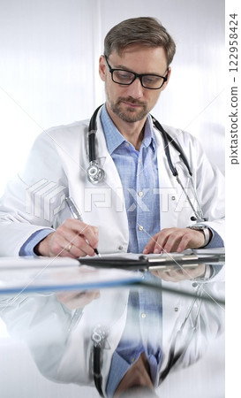 Doctor man in a blue shirt under a medical coat is using a clipboard and taking notes on glass desk in a medical office, vertical portrait view. Medicine and science Doctor man in a blue shirt under a medical coat is using a clipboard and taking notes on glass desk in a medical office, vertical portrait view. Medicine and science 122958424