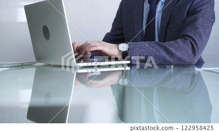 Businessman in blue jacket is focused on typing on laptop at glass desk in modern office, showcasing productivity and concentration, closeup view. Business people concept 122958425