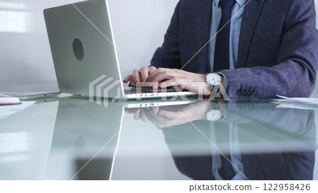 Businessman in blue jacket is focused on typing on laptop at glass desk in modern office, showcasing productivity and concentration, closeup view. Business people concept Businessman in blue jacket is focused on typing on laptop at glass desk in modern office, showcasing productivity and concentration, closeup view. Business people concept 122958426