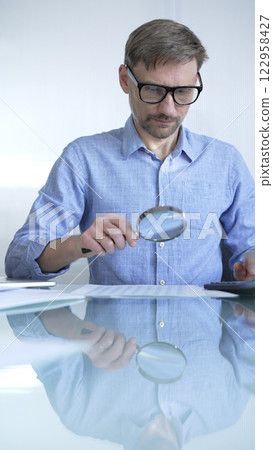 Businessman or accountant, wearing blue casual shirt and glasses, is using magnifying glass and calculator to analyze financial documents in office setting. Audit and taxes in business 122958427