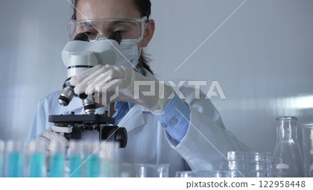 Scientist woman, wearing a lab coat, mask, white gloves and safety glasses, is using a microscope surrounded by test tubes in laboratory. Portrait view. Medicine and science concept 122958448