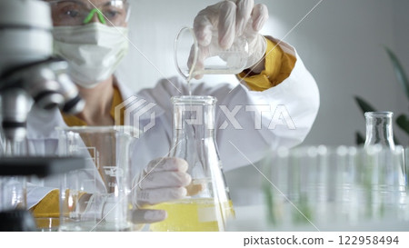 Woman scientist wearing a lab coat, white gloves, and a mask is pouring a yellow oily liquid from a beaker into an Erlenmeyer flask in a laboratory setting, close up. Science and medicine 122958494