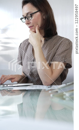 Focused businesswoman in brawn dotted blouse is using laptop and thinking with hand on chin, working on a project in a bright office. Business people concept Focused businesswoman in brawn dotted blouse is using laptop and thinking with hand on chin, working on a project in a bright office. Business people concept 122958497