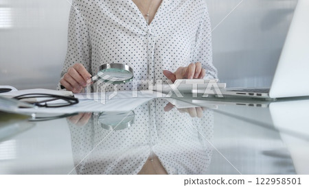Businesswoman carefully reviewing financial reports with magnifying glass and calculator, conducting audit and analyzing data, closeup view. Taxes and audit in business 122958501