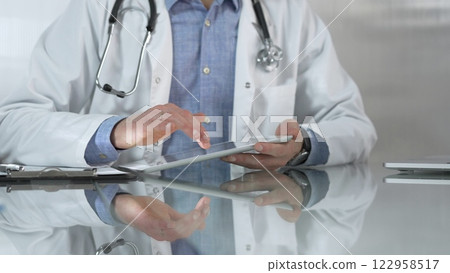 Doctor man, wearing a blue shirt under a medical white coat, is using digital tablet in modern healthcare setting, interacting with patient data, close-up. Medicine and health care Doctor man, wearing a blue shirt under a medical white coat, is using digital tablet in modern healthcare setting, interacting with patient data, close-up. Medicine and health care 122958517